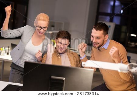 business, success and teamwork concept - happy coworkers with computer celebrating victory at night office