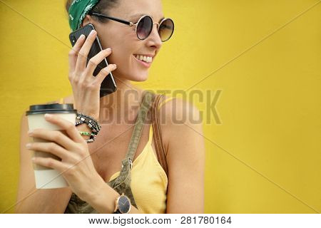 Styllish young woman on cellphone holding takeaway coffe outdoors on colored background