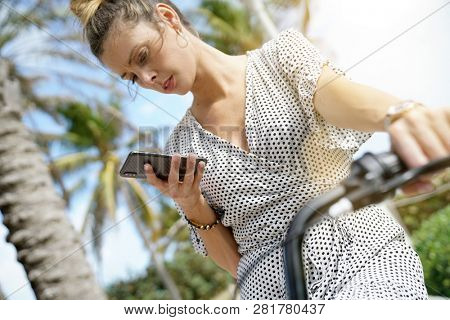 Pretty young woman sitting outdoors on bicylce reading directions on cellphone