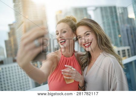 Two attractive funloving friends taking selfie on night out at roofttop bar in city