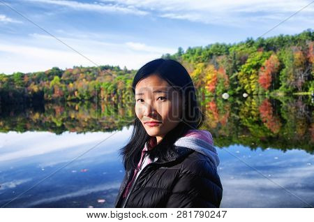 A Beautiful Chinese Woman Looking At The Camera With A Fall Foliage Reflecting In The Water Backgrou