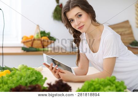 Young  Woman Looking For A New Recipe For  Cooking In A Kitchen. Housewife Is Making Online Shopping