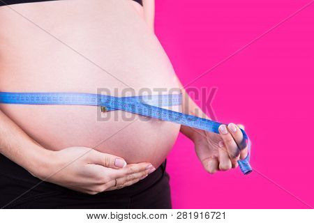 Close-up Belly Of Pregnant Woman With Tape Measure Tied Around On Pink Background. Measuring Circumf