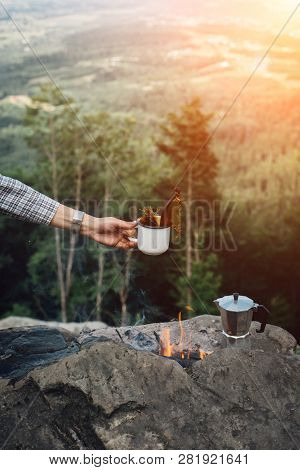 Girls Hand Holds A Hot Cup Of Coffee Near The Campfire On Background Of Nature During The Sunset . C