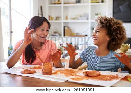 Two girlfriends having fun playing with modelling clay at home, laughing and showing their dirty hands to each other, close up