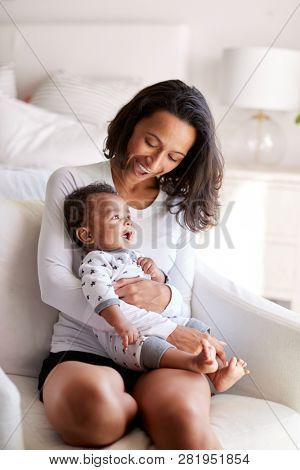 Young adult mother sitting in an armchair in her bedroom, holding her three month old baby son in her arms and looking down at him smiling, vertical