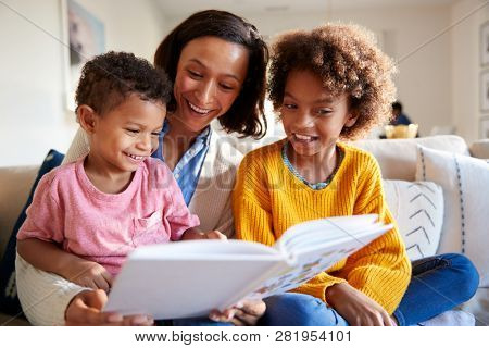 Close up of young mother sitting on sofa in the living room with her children, reading them a book, low angle