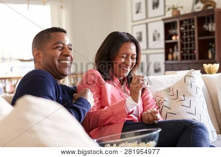 Close up of middle aged couple sitting on the sofa in their living room watching TV, laughing and eating popcorn, close up