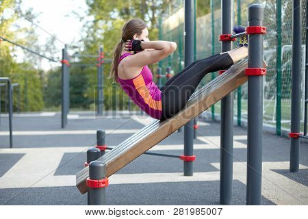 Young Caucasian Woman Workouts On The Park Sports Ground. The Girl Does Abdominal Excersise, In Blac