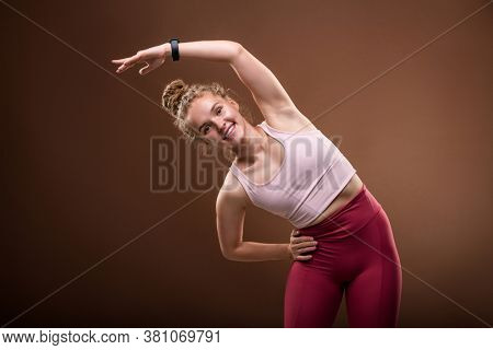 Cheerful young female in sportswear standing in front of camera and doing sidebends while enjoying morning workout against brown background