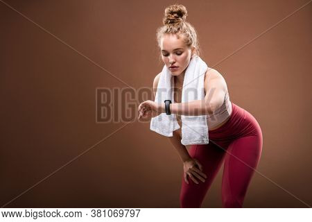 Tired young blond sportswoman in activewear looking at fitbit on her wrist while doing difficult physical exercise against brown background