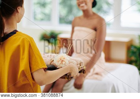 Masseur Bringing Tray With Hot Rolled Towels To Smiling Young Woman Sitting On Bed In Spa Salon