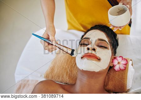 Cosmetologist Using Brush When Applying Mattifying Mud Mask On Face Of Pretty Young Woman