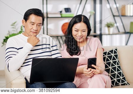Mature Asian Man Workng On Laptop When His Wife Sitting Near By And Watching Videos On Smartphone