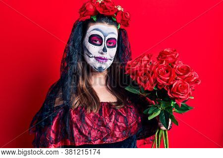 Young woman wearing day of the dead costume holding flowers looking positive and happy standing and smiling with a confident smile showing teeth 