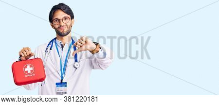 Handsome hispanic man wearing doctor coat holding first aid kit with angry face, negative sign showing dislike with thumbs down, rejection concept 