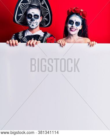 Young couple wearing mexican day of the dead costume holding blank empty banner looking positive and happy standing and smiling with a confident smile showing teeth 