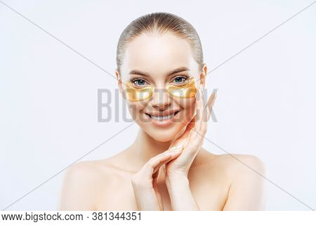 Studio Shot Of Attractive European Woman Touches Face Gently, Smiles Tenderly, Applies Golden Hydrog