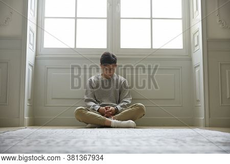 Young Asian Man Sitting On Floor At Home Head Down Legs Crossed