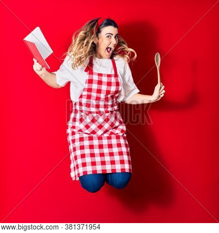 Young beautiful blonde cook woman wearing apron smiling happy. Jumping with smile on face holding recipe book and spoon over isolated red background