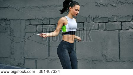 Horizontal Shot Of Athelete Determined Woman Skipping With Rope Outdoors Next The Cocnrete Wall. Fit