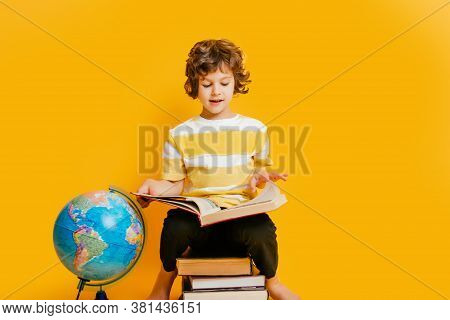 Curly Boy In Glasses And Stripes Tshirt And Sits On A Stack Of Books, Near To The Globus On Yellow B