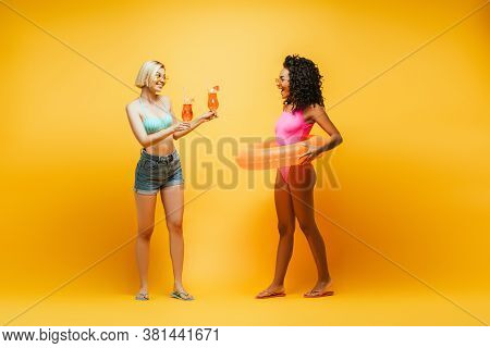 Blonde Woman Holding Cocktail Glasses Near African American Friend With Swm Ring On Yellow