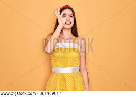 Young beautiful pin up woman wearing 50s fashion vintage dress over yellow background doing ok gesture with hand smiling, eye looking through fingers with happy face.