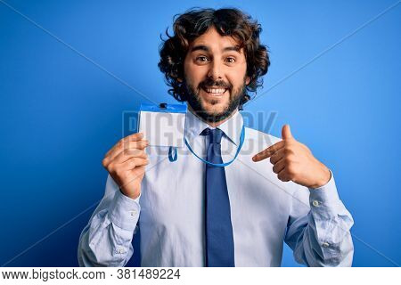 Young handsome business man with beard holding id card identification over blue background with surprise face pointing finger to himself