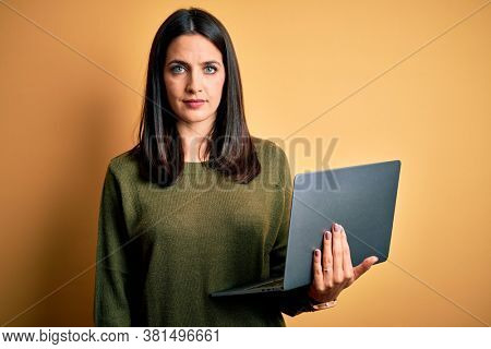 Young brunette woman with blue eyes working using computer laptop over yellow background Relaxed with serious expression on face. Simple and natural looking at the camera.