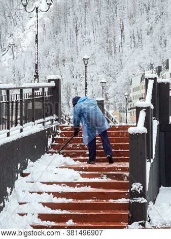A Man In A Plastic Raincoat Removes Snow On The Steps Against The Background Of Snow-covered Trees