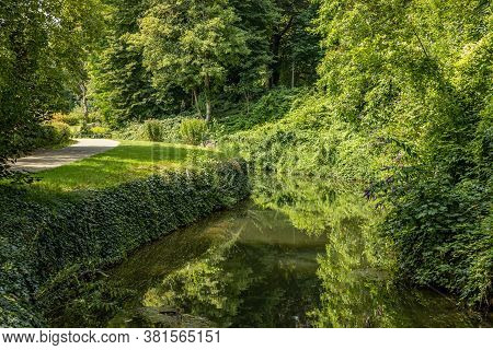 Jeker River Next A Path In The Maastricht City Park, Surrounded By Trees And Abundant Green Vegetati