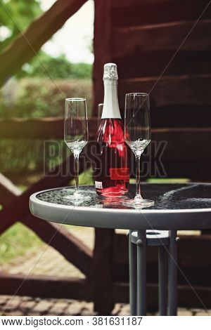 Two Flute Glasses And An Pink Champagne Bottle With No Label On A Glass Table In A Back Yard.