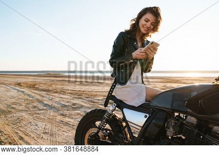 Beautiful young woman wearing lether jacket sitting on a motorbike at the beach, using mobile phone