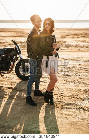 Beautiful young cople wearing jackets spending time at the sunny beach, standing near a motorbike, hugging