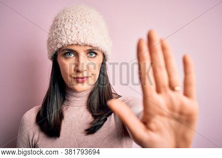 Young brunette woman with blue eyes wearing casual t-shirt and wool cap standing with open hand doing stop sign with serious and confident expression, defense gesture