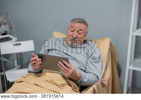 Medicine, Age, Health Care And People Concept - A Senior Man With A Tablet In Bed Of A Nurcing Home