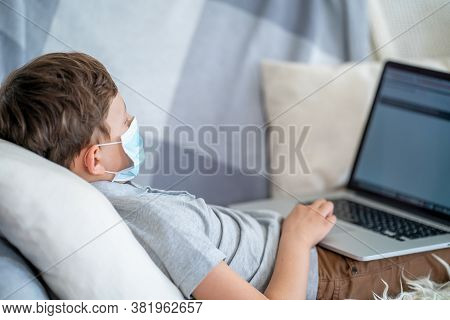 Focused Boy In A Mask, Lying On The Couch With A Laptop During The Quarantine Period. He Does Not Go