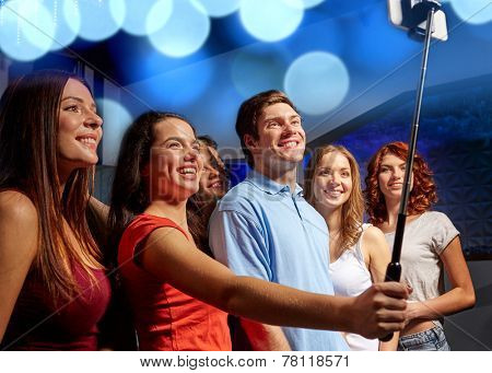 party, technology, nightlife and people concept - smiling friends with smartphone and monopod taking selfie in club