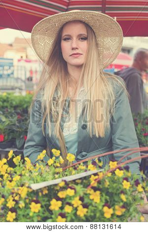 Attractive blonde girl with straw hat posing on flower marketplace. Post processed with vintage filter.