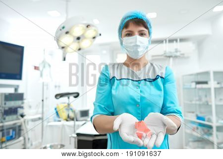 Shot of s female doctor wearing surgical mask and sterile medical gloves holding pink ribbon breast cancer awareness symbol posing in an operating theatre healthcare medicine mammalogy concept.