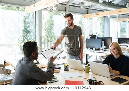 Image of smiling young colleagues sitting in office coworking using laptop computers and work with diagrama. Looking aside.
