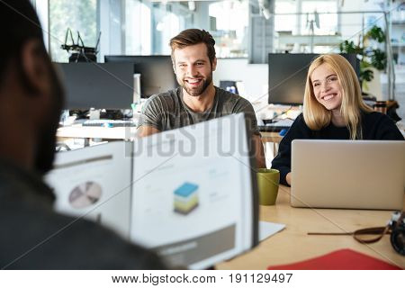 Picture of happy young colleagues sitting in office coworking using laptop computers and holding diagrama. Looking aside.
