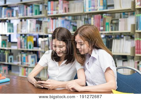 Happy two young Asian female students sitting at table and reading text message in libraly in university.