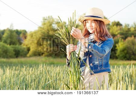 Happy young girl in summer hat collecting stems on wheat field. Summertime outdoors.
