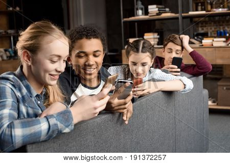 Happy Multicultural Teenagers Group Using Smartphones And Sitting On Sofa At Home, Teenagers Having