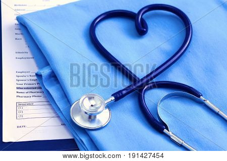 Medical stethoscope twisted in heart shape lying on patient medical history list and blue doctor uniform closeup.