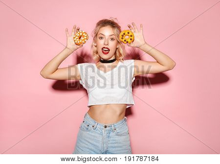Young fit hipster female in casual clothing holding doughnuts and looking at camera on pink background.