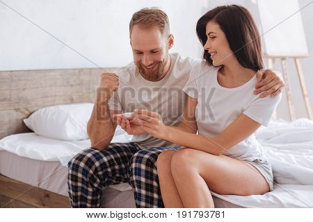 In seventh heaven. Extremely happy couple sitting on a bed and hugging while finding out results of a pregnancy test.