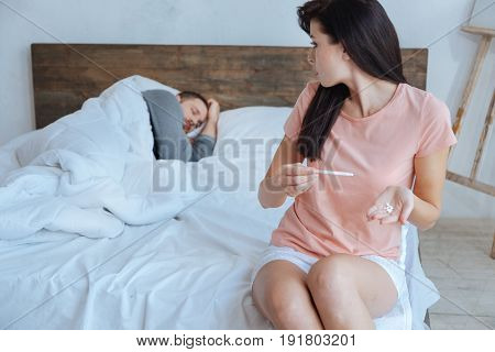 Maybe I should ask him. Young puzzled brunette looking at her boyfriend sleeping while holding a positive pregnancy test and a handful of pillows.